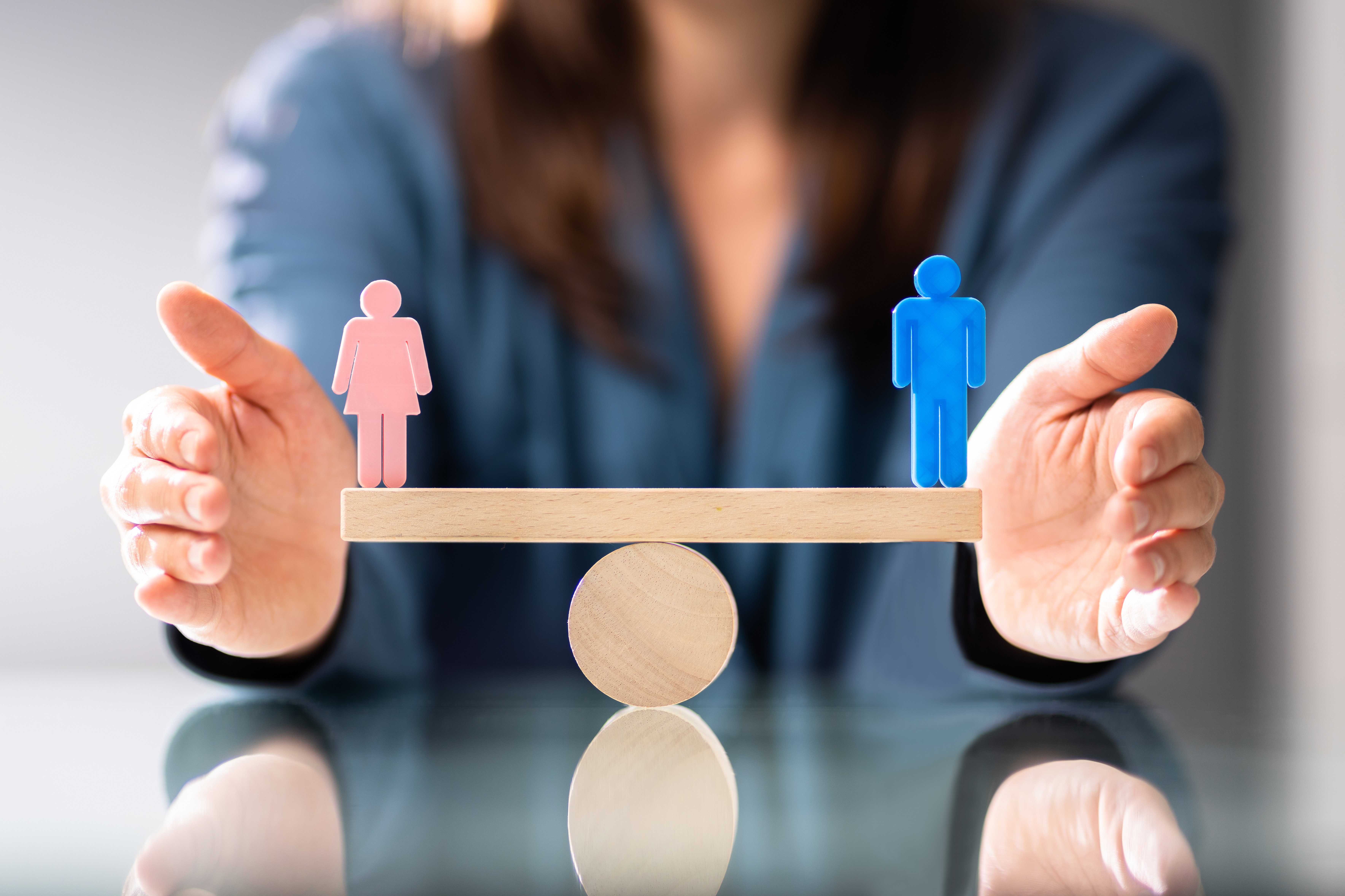 A wooden scale with a wooden man on one end, and a wooden woman on the other. The scales are perfectly balanced.
