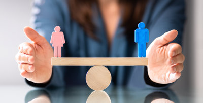 A wooden scale with a wooden man on one end, and a wooden woman on the other. The scales are perfectly balanced.